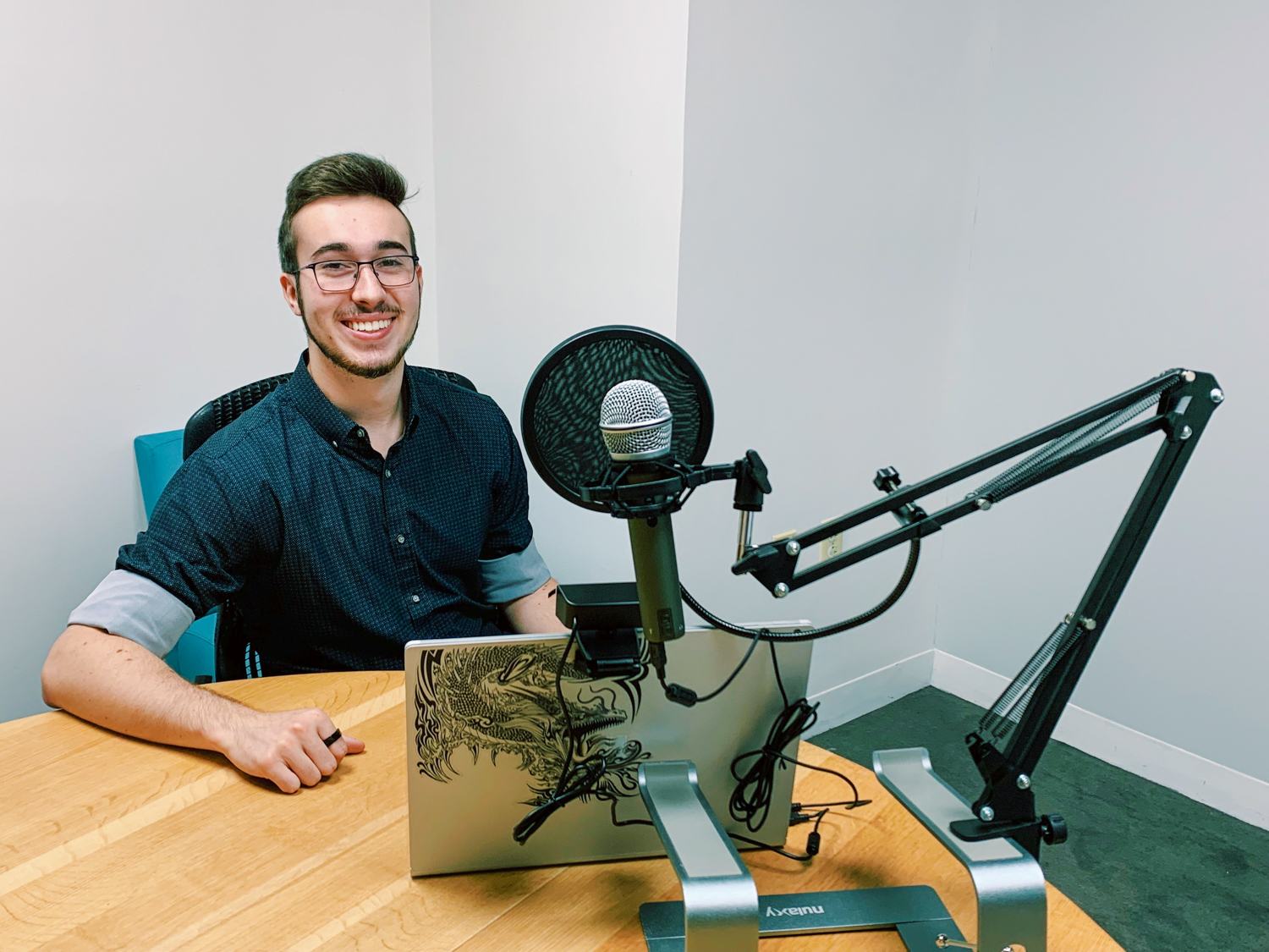 A male student seated at a table poses with his laptop and podcasting equipment