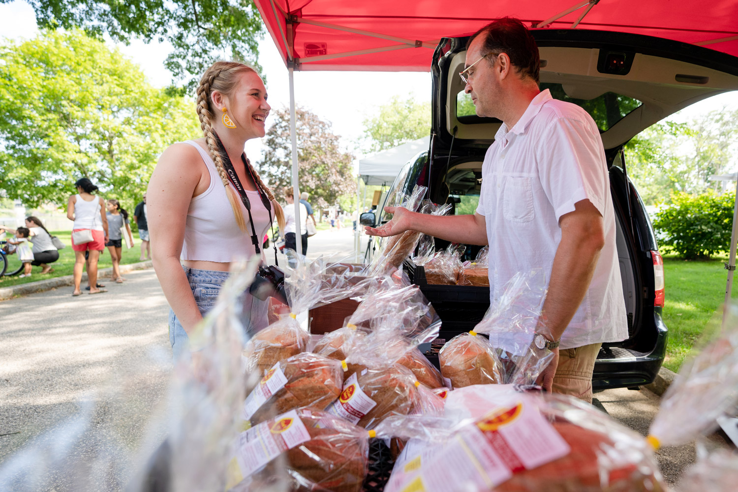 A female student stands at a table of bread at an outdoor farmer's market talking to an older man.