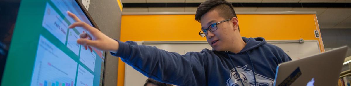 Student holding a computer in one hand and pointing to a screen in the other