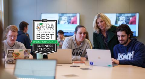 Inside a Paul College classroom, a female professor leans between two students to chat about a project shown on a laptop. A graphic for the Poets&Quants badge for Best Undergraduate Business School 2024 is placed to the left side.