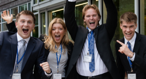 Three professionally dressed male students and one female student jump in the air outside, with Paul College behind them. Their expressions are jubilant and one of them is holding a glass award.