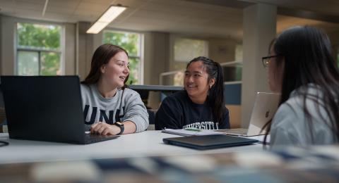 Three Paul College students sit at a table, two with open laptops and UNH apparel. In the background are windows with soft lighting streaming in through trees.