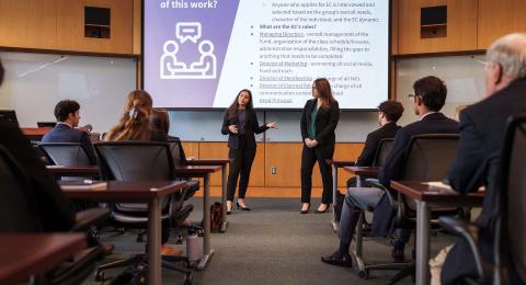 Two professionally-dressed female students gesture to a projector screen. In the mid-and foreground students and faculty sit at desks and watch the presentation.