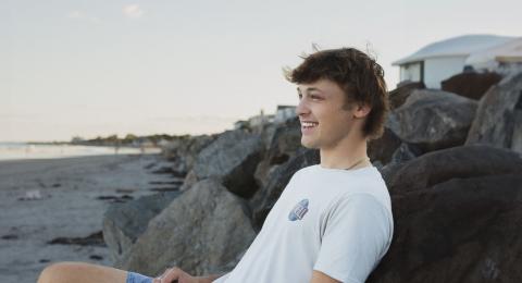 UNH Marketing and Finance Major Gavin Cieplik leans against some rocks on a local beach, smiling at something off camera.