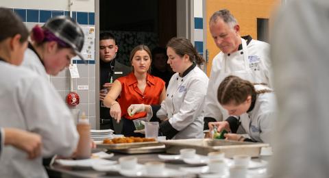 Four students in chef's coats are flanked around a metal prep table, one is putting the finishing touches on a dish, while the head chef and professor overlooks. In the background one professional dressed student in entering the kitchen through a doorway, with another student just behind her.