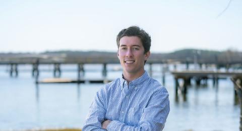 Paul College student stands with arms crossed, smiling. He wears a stripped button down & dress pants. In the background, a boat on land, and piers in the water.