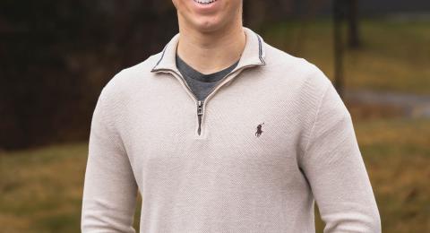 Accounting major Harrison smiles, standing on campus wearing an off-white quarter zip sweater. In the background, light green grass and a darker treescape.