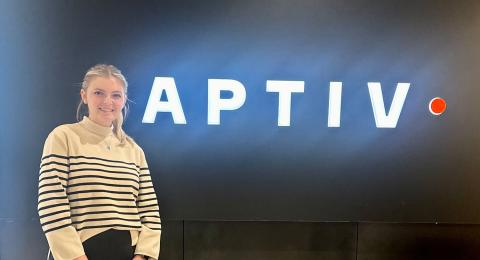 UNH Finance and Entrepreneurial Studies Major Lauren Chance stands next to the Aptiv sign in the lobby of her internship's office building, smiling at the camera.