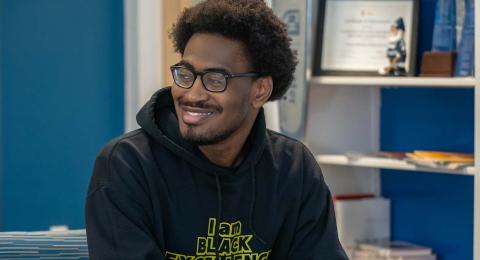 UNH Economics major Christian Katumba sits in a chair at the UNH ECenter, smiling at something off camera. He has an afro pick in his hair and is wearing a sweatshirt that says, "I am Black excellence."