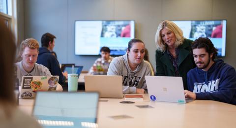 A UNH Paul College professor leans between two students working on a laptop during a business course in the collaborative classroom space.