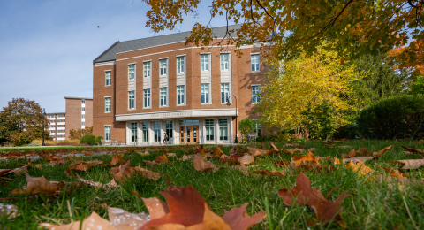Exterior of Paul College; a red brick academic building with white columns, photographed through autumn leaves on grass with fall trees framing the scene.