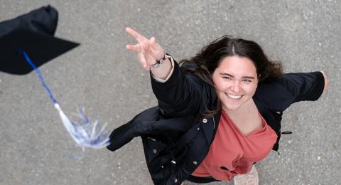 Female student throws a graduation cap into the air towards the camera, the tassle trailing. She has dark hair, and wears a coral shirt and black jacket.