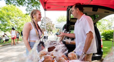 A female student stands at a table of bread at an outdoor farmer's market talking to an older man.