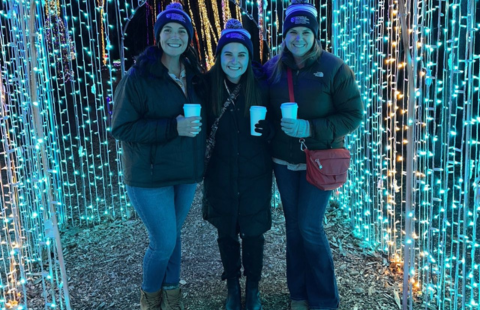 Annie Nevells, Sarah Hills, and Becky Barbour pose with coffee cups in handles, and bundled up in winter jackets and hats, in front of a stringed, light display outdoors