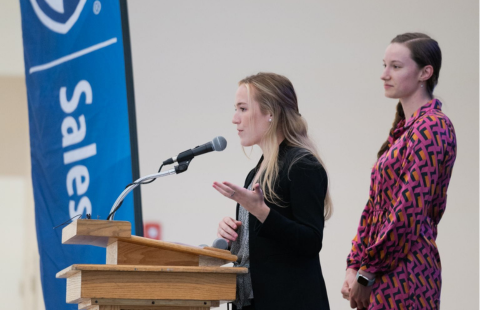 Two female students stand at a podium, one talking into the speaker, with a flag banner in the background showing the UNH logo and the word "Sales"