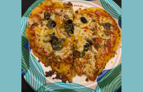 A small, heart shaped pizza with mutilple toppings sit on a paper plate with teal, and green designs.