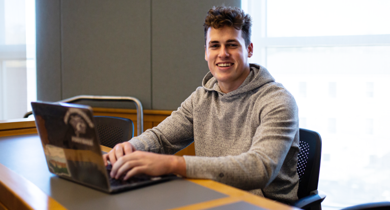 A male student sits in a classroom with his laptop in front of him, smiling at the camera