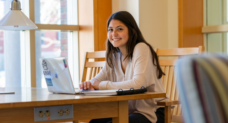 A female student sits at a table in the UNH library with her laptop and backpack, smiling at the camera.