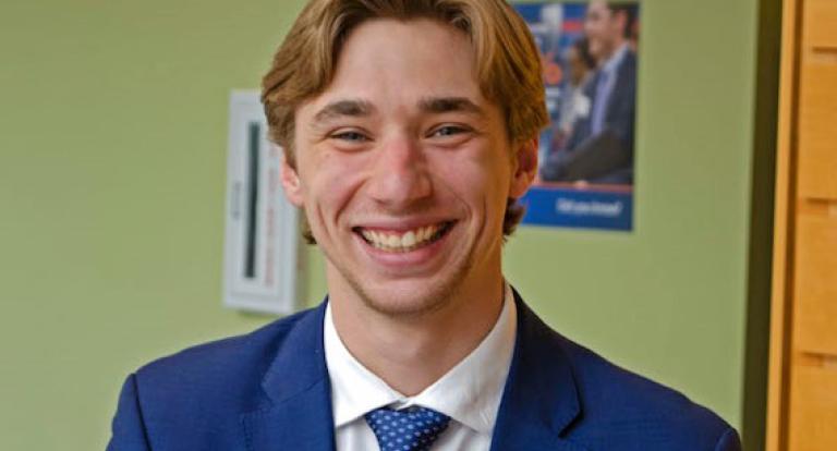 A male student wearing business attire smiles at the camera.
