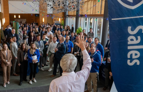 In the foreground, a back of a white-haired gentleman's head next to a flagged banner that reads "UNH Sales". He is waving to a mass of students in the background, who are dressed professionally and gathered in the brightly and natural lit Great Hall.