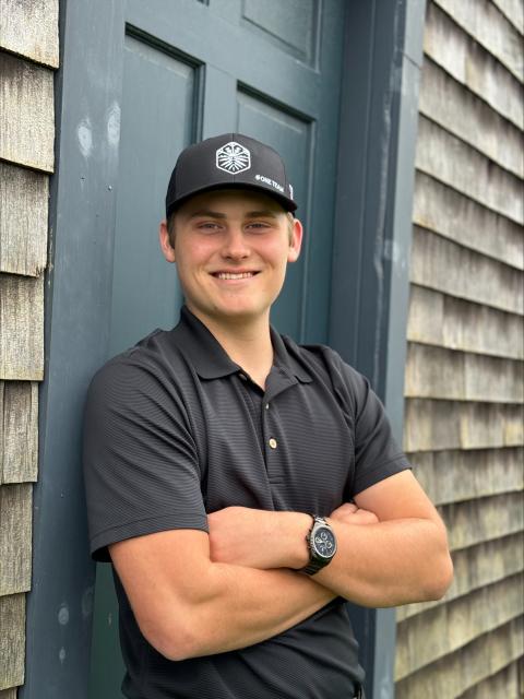 UNH Paul College student smiles with his arms crossed wearing a black polo and baseball cap. He leans against a doorframe of a house with dingy shingles.