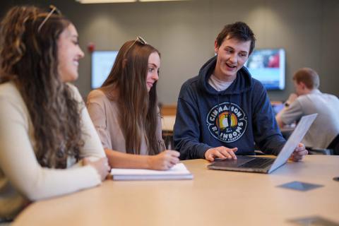 Three students from the Marketing and Advertising Club sit at a table in a classroom looking at a laptop together at UNH Paul College.