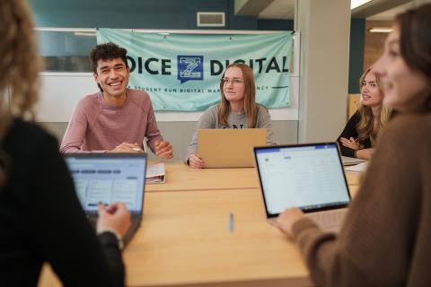 Five Voice Z Digital Marketing Agency students gather at a table in UNH Paul College's experiential learning space, the Forge, working on laptops and laughing.