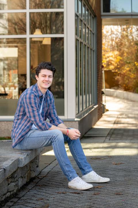 Analytical economics and student-designed major Sam Croteau sits outside the UNH Paul College building on the edge of a flowerbed, smiling at the camera.