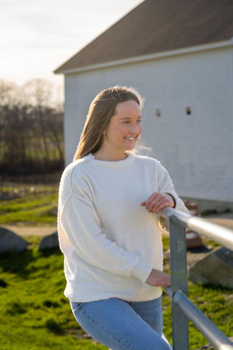 UNH accounting and finance major Reagan Calcari leans against a fence near the campus horse barns, smiling to someone off camera.