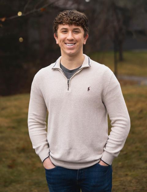 Accounting major Harrison smiles, standing on campus wearing an off-white quarter zip sweater. In the background, light green grass and a darker treescape.