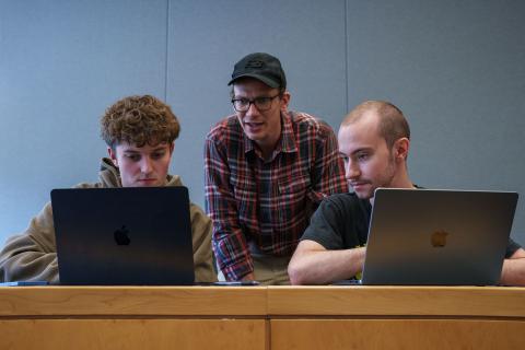 A teacher leans between two UNH students with laptops open in front of them to chat about what they're working on.