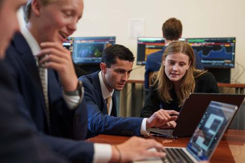 Students gather in the Bloomberg Terminal room in Paul College, with the camera focused on a male and female student looking over something on a laptop together.