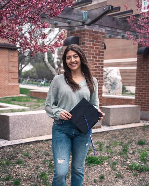 UNH Economics major Emily Bolognino stands on UNH campus in front of a blossoming tree, smiling at the camera, holding a graduation cap.