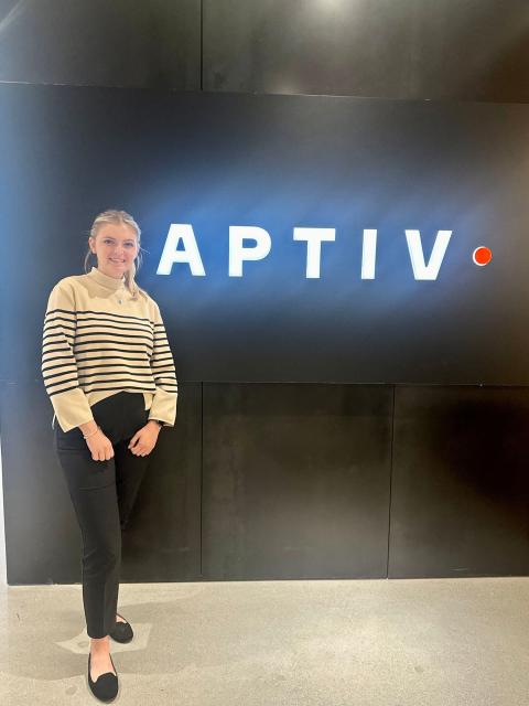UNH Finance and Entrepreneurial Studies Major Lauren Chance stands next to the Aptiv sign in the lobby of her internship's office building, smiling at the camera.