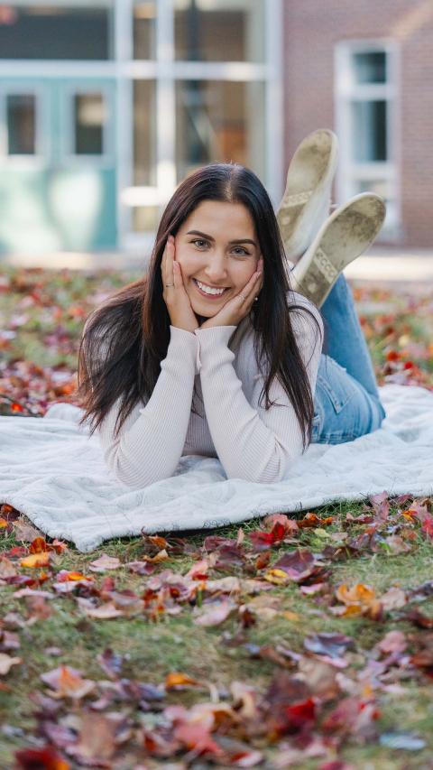 UNH International Business and Economics major and transfer student Gabriella Caterino lies on a blanket on top of an autumn-leaf strewn lawn, hand under her chin and legs kicked up behind her in the air, grinning at the camera.