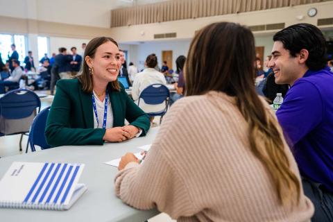 A female student sits at a table in UNH's memorial union building and smiles at two professionals seated in front of her during UNH Sales Center's annual Speed Sell event.