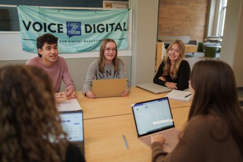 Members of UNH Paul College's student-run digital marketing agency, Voice Z, gather at a table with their laptops, a banner that reads "Voice Z Digital" hanging behind them.