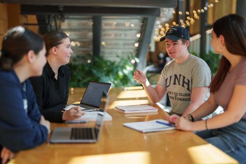 A group of students sit and chat at a table in UNH Paul College's Great Hall.