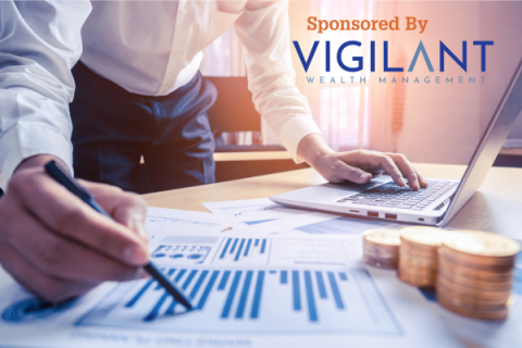 Professionally dressed man stand with pen in hand atop reports on a desk, cold coins are stacked the right. Overlayed, "Sponsored by Vigilant Wealth Management"