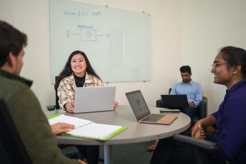 Graduate students enrolled at UNH Peter T. Paul College of Business and Economics gather in a graduate study space inside the building, chatting at a table with their laptops.