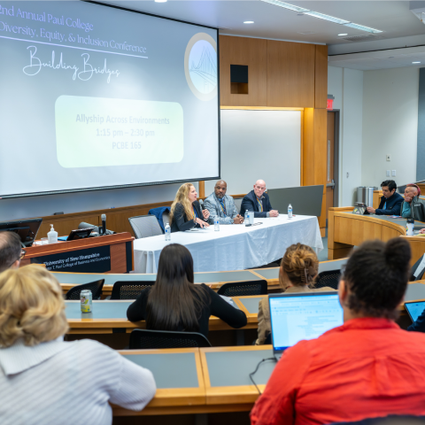 Participants sit in a classroom facing the front of the room where three panelist sit with a projector screen behind them