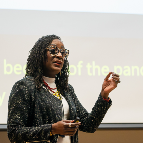 Woman with dark hair and glasses stands in front of a project screen and gestures with her hands as she gives a lecture