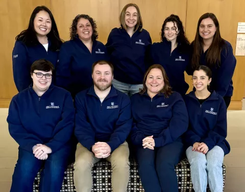 Eight female staff and one man staffer from the UNH undergrad team pose for a group photo wearing matching quarter zip sweatshirts. Half are seated.