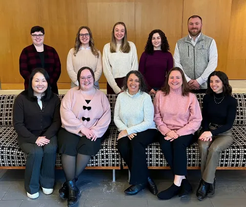 : Group of ten staff members seated and standing on a patterned bench against a wood-paneled wall, wearing winter sweaters