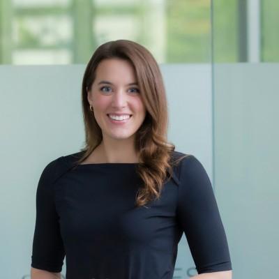 Professional headshot of Alexys Febonio with shoulder-length hair, wearing a black top, smiling, with a softly lit office background.