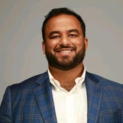 Professional headshot of Hanzla Sheikh with short hair and a beard, wearing a blue plaid blazer and white shirt, against a gray background.