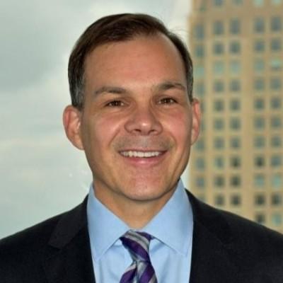 Professional headshot of  Mark Carver in a suit and striped tie, smiling, with a blurred city building in the background.