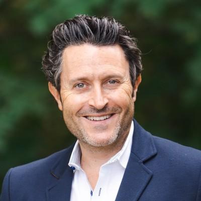Professional headshot of Rick Marini in a navy blazer and white shirt, smiling, with a blurred green outdoor background.