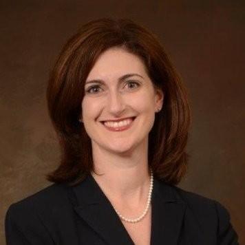 Professional headshot of Meg Rotondo in a dark blazer and pearl necklace against a brown studio background.