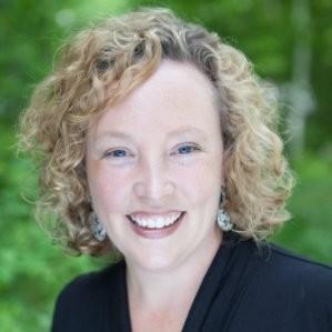 Head-and-shoulders portrait of a Amy McLaughlin with curly light hair, wearing a black top against a softly blurred green background.